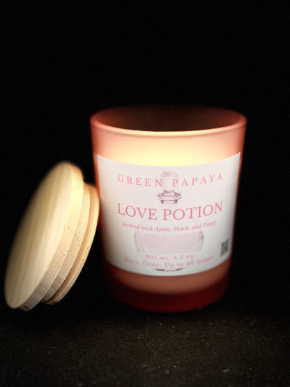 A lit candle in a frosted peach-colored glass jar with a white label reading 'GREEN PAPAYA LOVE POTION,' shown on a black background with a light wood lid leaning beside it.