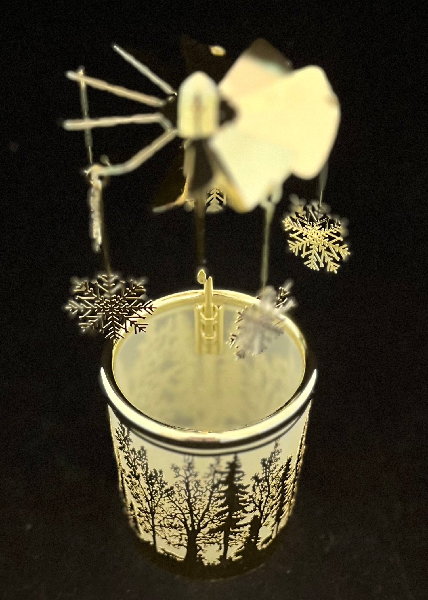 A decorative winter-themed candle holder featuring a white and black forest tree patterned cup with a wind-up spinner top and hanging snowflakes on a black background.