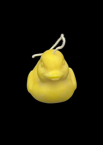 A small yellow duck-shaped candle with a white wick, photographed against a dark black background.